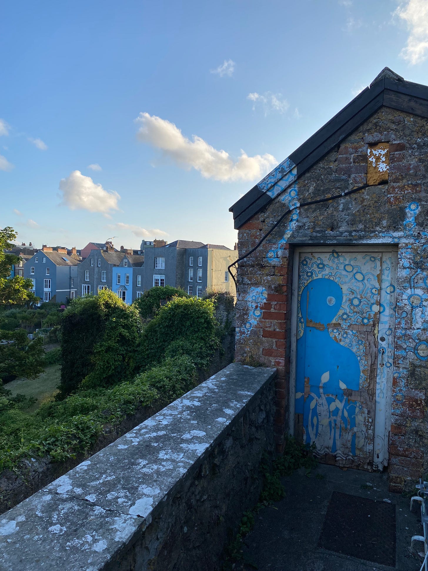 a brick building inn the foreground with a door covered in a blue images the same colour as the sky which hags above distant grey buildings with white window and door frames separated from the viewer by a green hedge