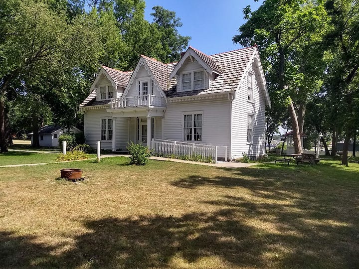 Modest one story house with attic rooms set in large lawn with cars and development glimpsable across the main road plus historic signs