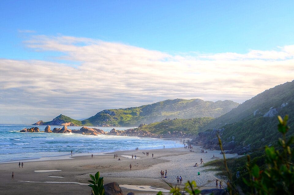 A wide coastal landscape of Praia da Galheta seen from an elevated trail. Soft late-afternoon light illuminates the sandy beach, scattered beachgoers, rocky outcrops, and green hills rising behind the shoreline. Waves roll in from the right under a sky filled with low clouds and blue patches. A wide coastal landscape of Praia da Galheta seen from an elevated trail. Soft late-afternoon light illuminates the sandy beach, scattered beachgoers, rocky outcrops, and green hills rising behind the shoreline. Waves roll in from the right under a sky filled with low clouds and blue patches.