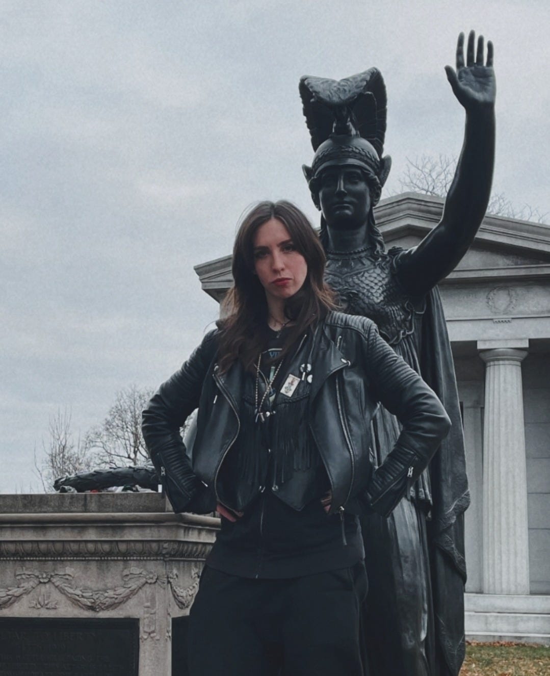 The author standing in front of a statue of the Roman goddess, Minerva, in Green-Wood Cemetery, in Brooklyn. The sky is grey, and Claymore, clad in black and a leather jacket, stares at camera, with hands on their hips triumphantly.