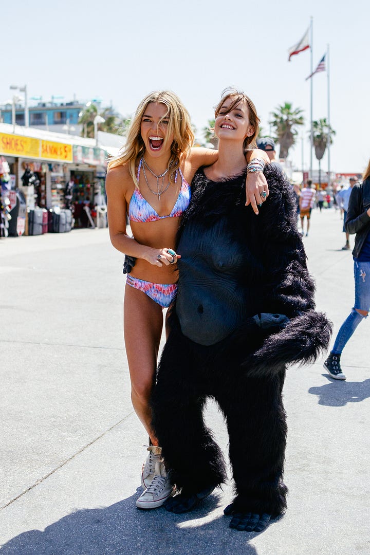 Barbara Palvin and Ashley Hart on set, laughing and posing with a gorilla suit—pulling it on, peeling it off, and mixing fashion with absurdity during the Mambo Goddess campaign shoot.