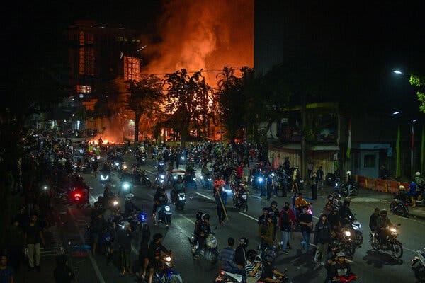 Dozens of demonstrators on a wide street in front of a building that is engulfed in flame.
