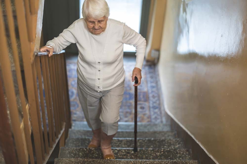 Elderly woman struggling to walk up the stairs while leaning on a wooden cane.