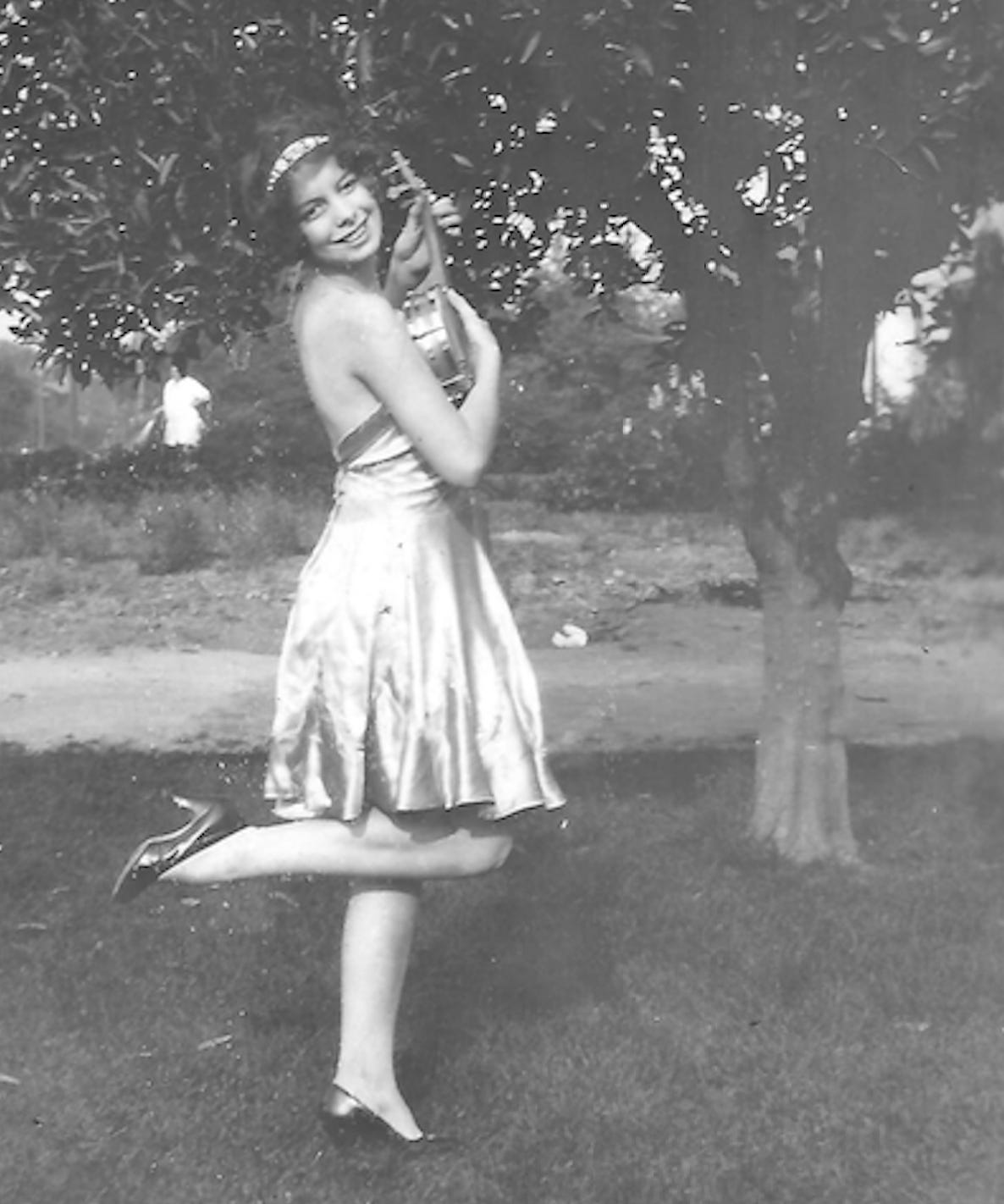 Sally Tamblyn stands outside on grass. She smiles at the camera. She is playing what appears to be a tiny banjo. She wears a headband in her hair and is dressed in a sleeveless, knee-length dress and high heels.