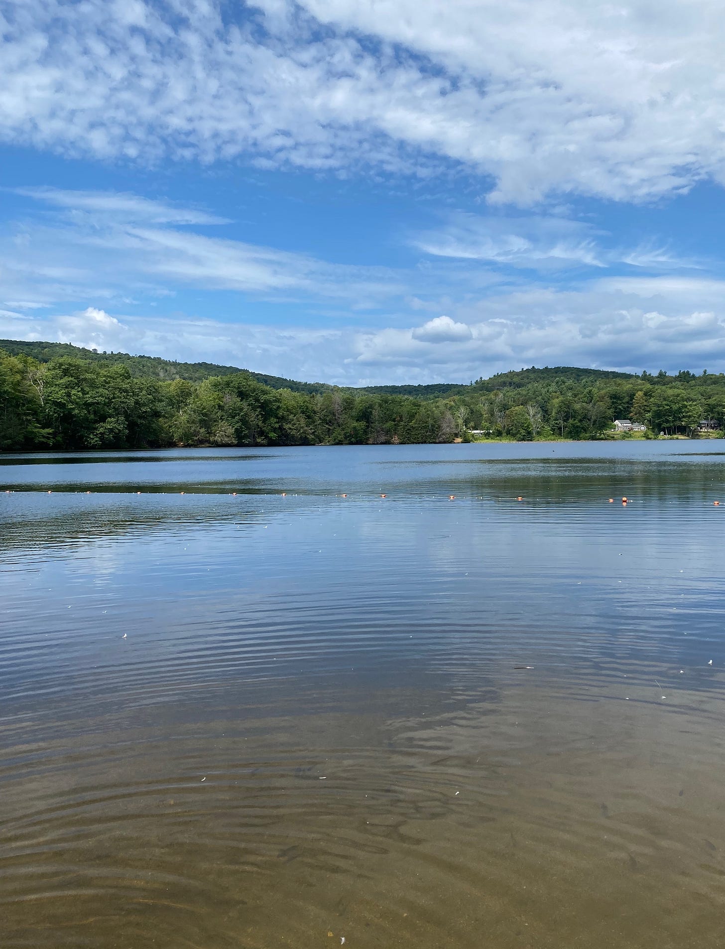 A clear lake, light ripples across its surface, surrounded by green hills and a blue, cloud-studded sky.