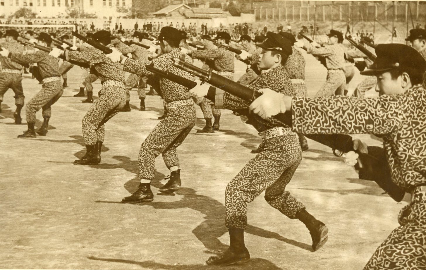 Rows of Korean high-school boys in patterned uniforms and caps practice rifle drill with training rifles on a dirt field during a 1972 gyoryeon (교련) military-training class in Gangwon Province; school buildings and onlookers in the background.