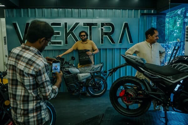 A man stands with a new motorcycle in front of a sign that reads “Vlektra” while someone snaps a photo of him with a cellphone.