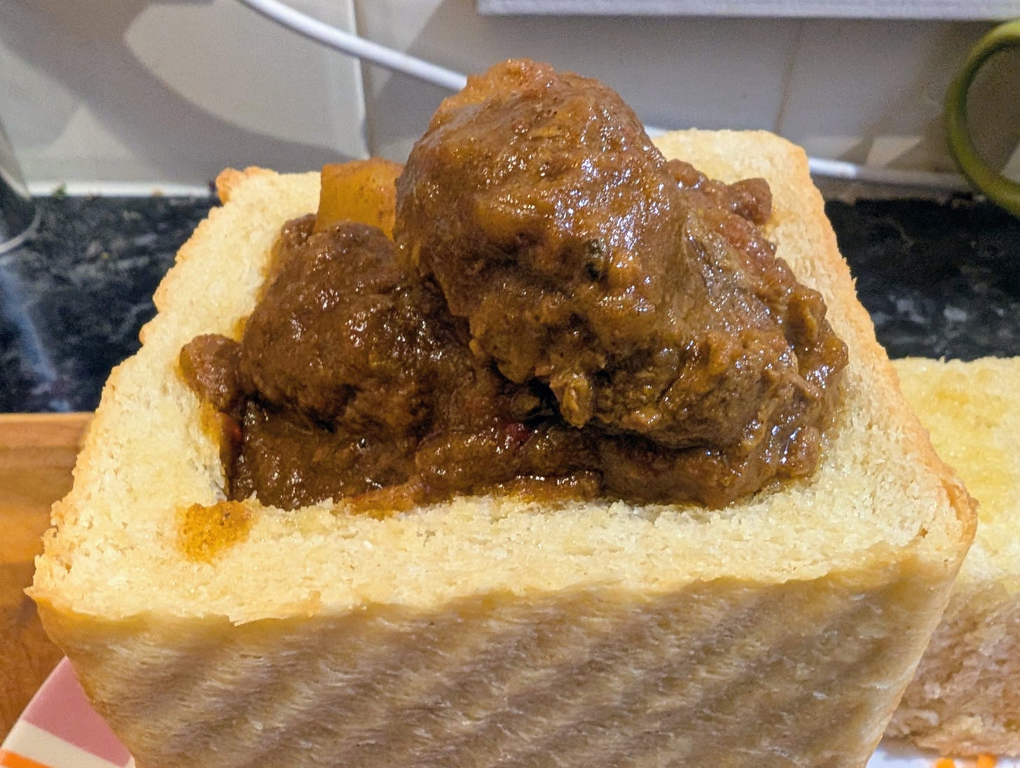 A hollowed-out square loaf of white bread is filled to the brim with a thick, brown South African curry containing chunks of meat and potato. The bread’s soft interior has been removed to form a container, and the curry sits glossy and dense inside it.