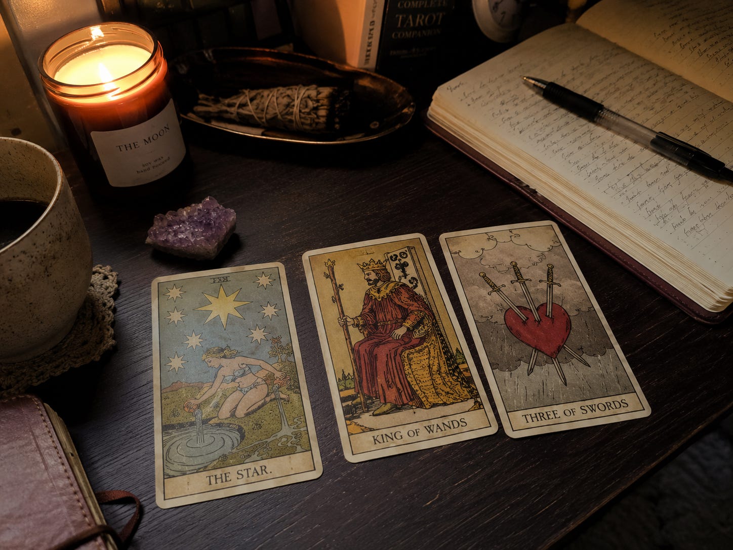 A three-card tarot spread laid out on a dark desk beside a lit candle, an open journal, and an amethyst crystal — The Star, King of Wands, and Three of Swords, left to right.A three-card tarot spread on a dark wooden table — The Star, King of Wands, and Three of Swords — beside a lit candle, an open journal, and an amethyst crystal
