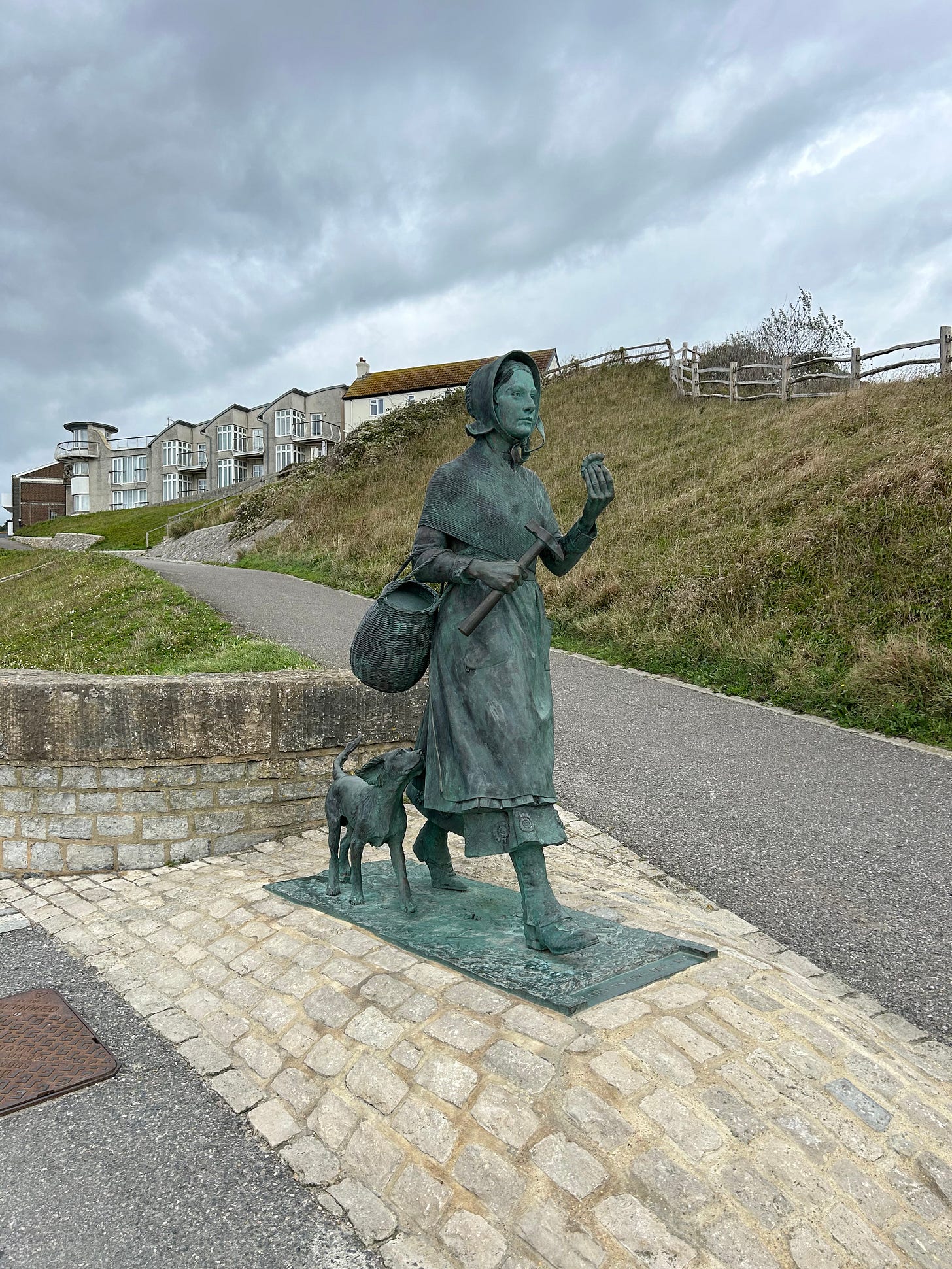A bronze statue of Mary Anning and her dog. She is looking out to sea across a bay not in view in this photo. In the background is a grassy bank and some houses on the horizon. Image: Roland’s Travels A bronze statue of Mary Anning and her dog. She is looking out to sea across a bay not in view in this photo. In the background is a grassy bank and some houses on the horizon. Image: Roland’s Travels