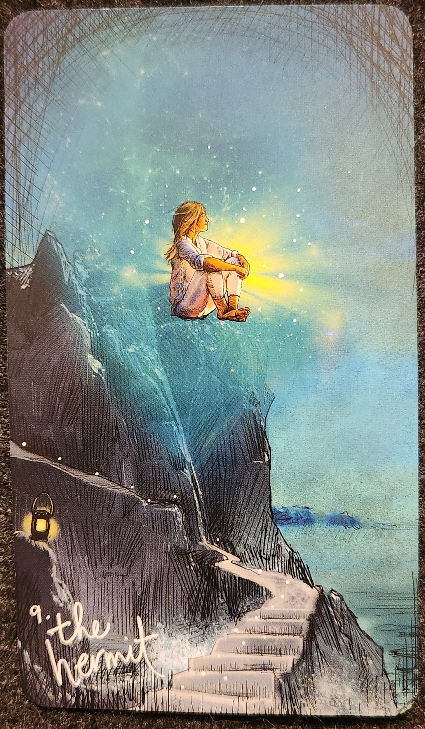 A tarot card labelled 9. The Hermit. A person sits hugging their legs at the top of a cliff. They are surrounded by a bright glowing light. Their eyes are closed and they are facing over the water, wind blowing their hair. The path to the cliff is a narrow set of stone stairs. There is a small lantern sitting on a rock at the bottom of the steps. 