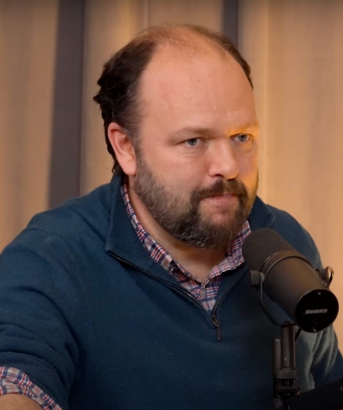 Ross Douthat sitting in front of a microphone; close up; he is white, bearded, somewhat balding, wearing a blue sweater Ross Douthat sitting in front of a microphone; close up; he is white, bearded, somewhat balding, wearing a blue sweater