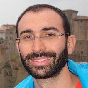 Retrato em plano fechado de um homem de cabelos curtos e escuros, óculos de grau sem aro e barba cheia, sorrindo com uma jaqueta azul e camiseta laranja.