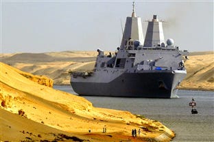 The color image is of the USS San Antonio, United States Navy Ship while transiting the Suez Canal. The ship is view from the starboard quarter bow on in the distance plying slowly in the canal. On both sides of the image are sand dunes giving the sense that the ship is in the canal.