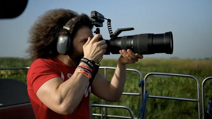 Photographer outdoors holding a camera with a long telephoto lens and a small device mounted on top via a coiled cable, wearing large headphones and a red shirt, shooting over a grassy landscape.