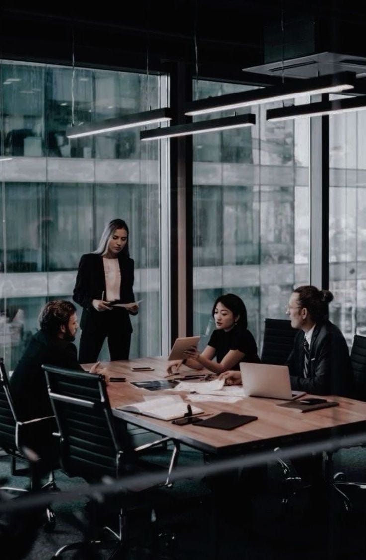 This may contain: a group of people sitting around a wooden table in an office setting with glass walls
