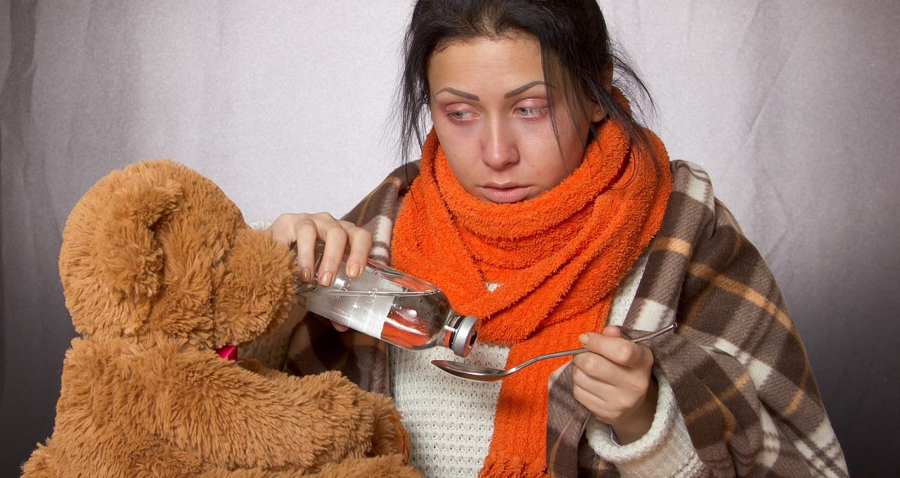 A sick woman pours some medicine onto a spoon for a teddy bear. A sick woman pours some medicine onto a spoon for a teddy bear.