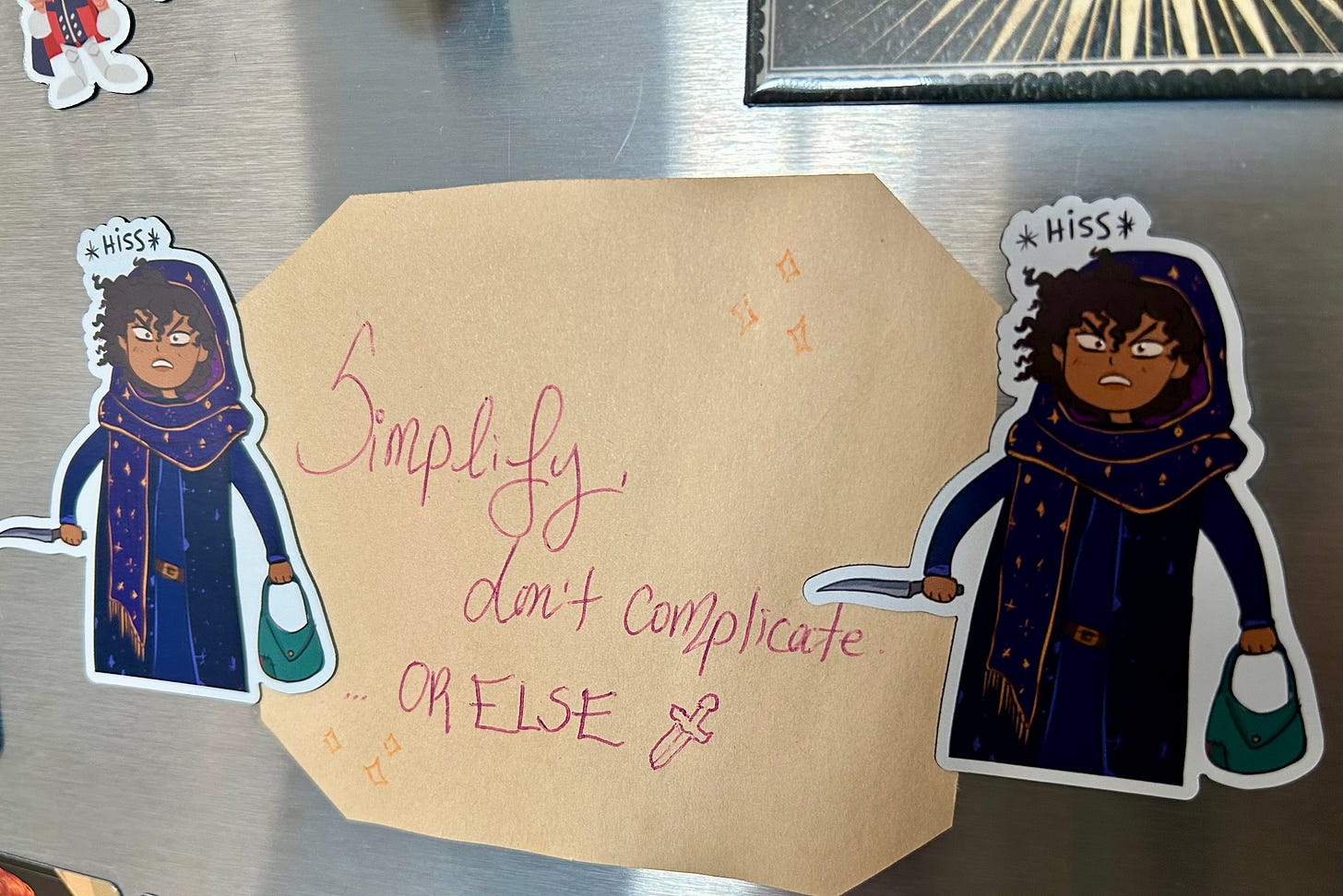 Two angry Loulie magnets hold up a piece of paper that reads: "Simplify, don't complicate. ...or else."