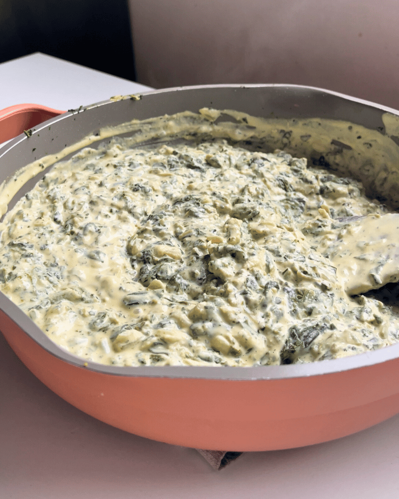 creamy spinach and artichoke filling in a pink pan with a wooden spatula stirring it