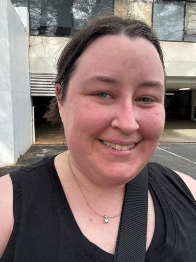 A selfie of a early 40s woman with a red face, standing in a parking lot. She's wearing a black tank and has her hair up