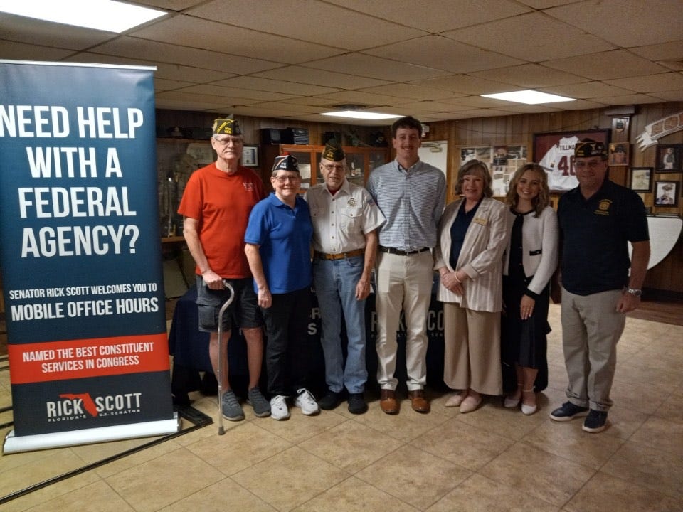 Seven adults stand side by side inside a wood-paneled community hall, some wearing military veterans’ caps, posing in front of a table and display. A large banner on the left reads: “NEED HELP WITH A FEDERAL AGENCY? SENATOR RICK SCOTT WELCOMES YOU TO MOBILE OFFICE HOURS. NAMED THE BEST CONSTITUENT SERVICES IN CONGRESS,” with “RICK SCOTT FLORIDA’S U.S. SENATOR” at the bottom. A table behind them also shows “RICK SCOTT.” The room has framed photos and a framed sports jersey on the wall; one person holds a cane.