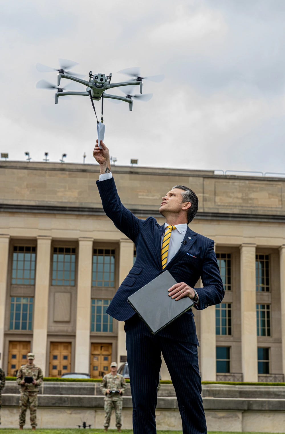 Color photo of Secretary of Defense Hegseth reaching up to receive a memo delivered by a small unmanned air vehicle (UAV) hovering just about his head. The UAV is a quadcopter approximately 2 feet in diameter. The photo backdrop is the pentagon with two military service members standing in the background dressed in camouflage uniforms, one of whom is operating the UAV.