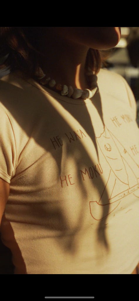 women's baby tee in tan with brown lettering and a drawing of a waʻa or Hawaiian style voyaging canoe. Wandering through the forest.