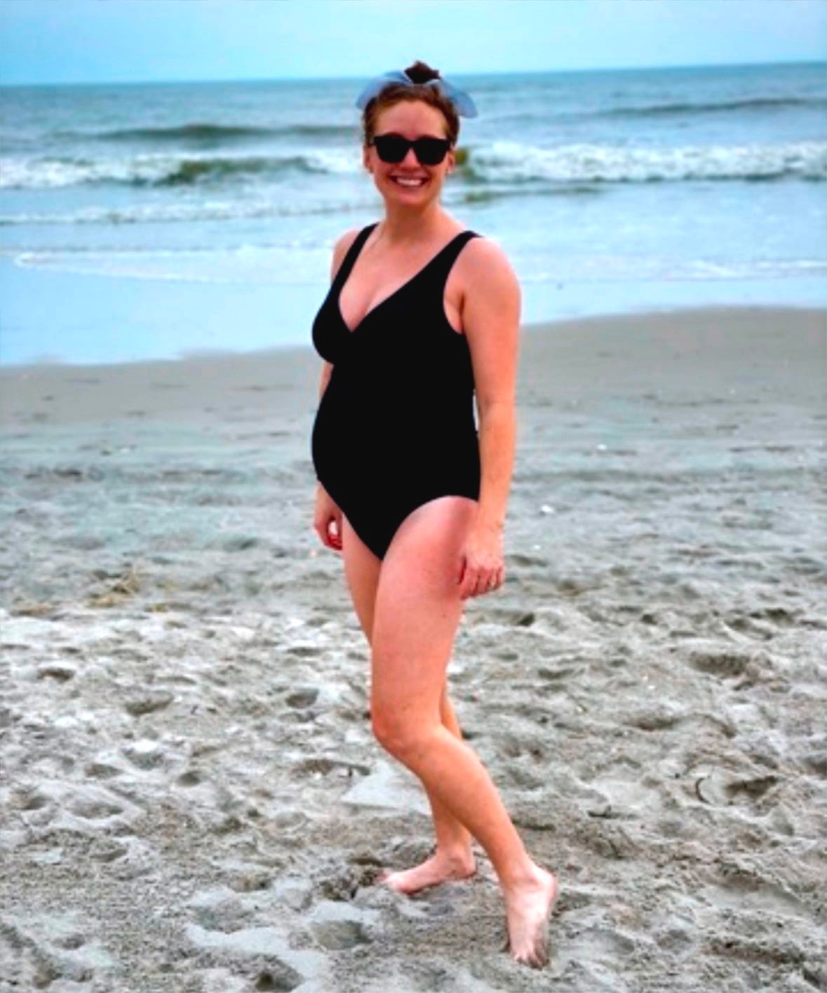 Angela standing on a beach while pregnant with twins, wearing a navy swimsuit and smiling, with waves and blue sky in the background. Angela standing on a beach while pregnant with twins, wearing a navy swimsuit and smiling, with waves and blue sky in the background.