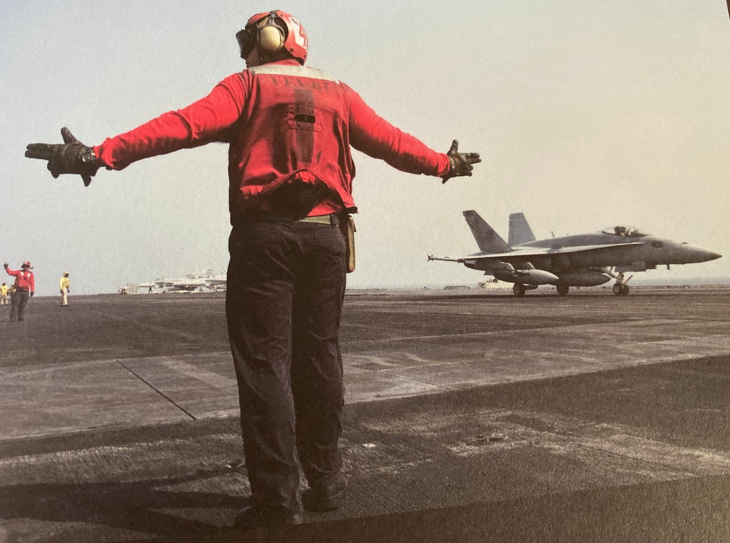 aircraft carrier with man in foreground and plane on the deck in middle ground taken from the book Another Great Day at Sea by Geoff Dyer 