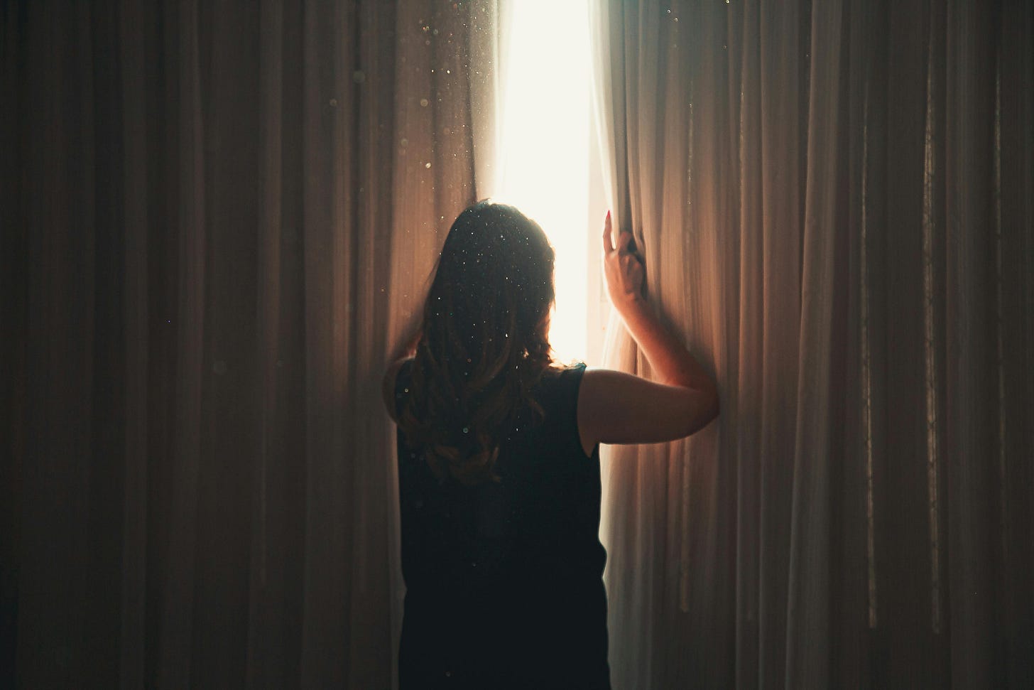 A imagem mostra uma pessoa vista de costas, com cabelos longos e ondulados, usando uma blusa sem mangas. Ela está abrindo uma cortina espessa, permitindo que a luz do sol entre no ambiente. A luz ilumina parcialmente a pessoa e cria um efeito visual bonito, destacando as partículas de poeira que estão suspensas no ar. O cenário sugere um momento tranquilo e contemplativo, como alguém explorando o que está além da cortina ou permitindo que a luz ilumine o espaço em volta. A composição da imagem evoca sentimentos de curiosidade, introspecção e renovação.
