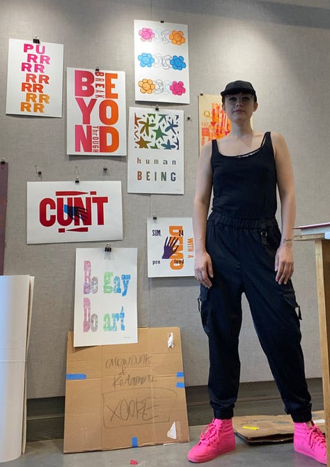 Shady standing in front of several of their letterpess prints at Penland, a letter pressprint reading "Beyond, Break Through" sitting on the press, and a print that says "Gay Art".