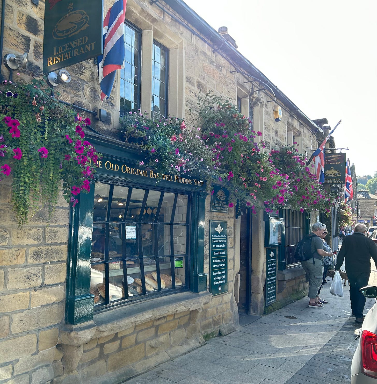 A photo of the double-fronted Original Bakewell Pudding Shop. People are walking by along the pavement. Flowers hang over the windows. A photo of the double-fronted Original Bakewell Pudding Shop. People are walking by along the pavement. Flowers hang over the windows.