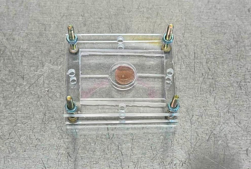 Cápsula de acrílico transparente com estrutura retangular contendo uma moeda de um centavo de dólar americano no centro, sustentada por suportes internos, com quatro parafusos de latão posicionados nos cantos e dois parafusos adicionais no topo, fotografada sobre uma superfície têxtil cinza.