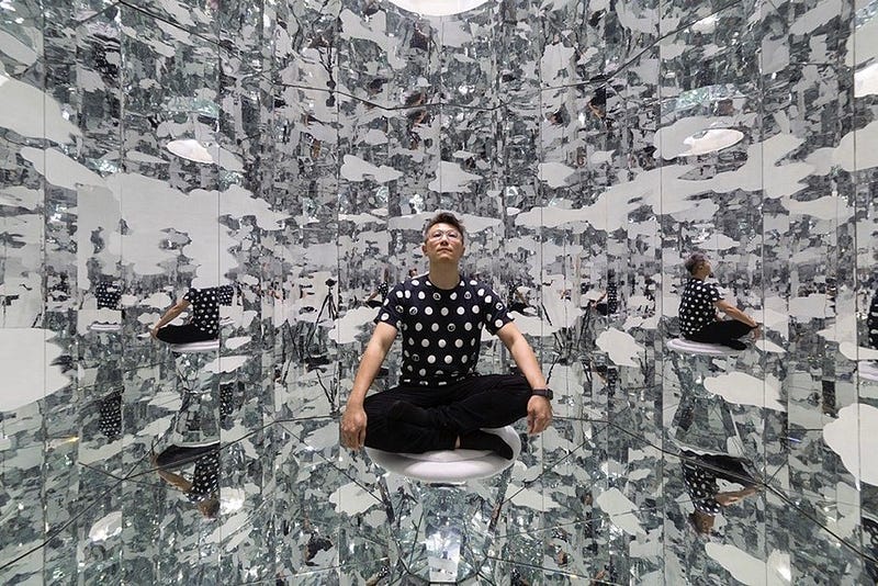 A man sits on a stool in the center of a completely mirrored chamber: the ceiling, floor, and walls are all mirrored and the chamber is rounded. It’s hard to know its exact shape and where its edges are. The man can be identified in some reflections, but there are also indistinguishable shapes that look like smeared clouds.
