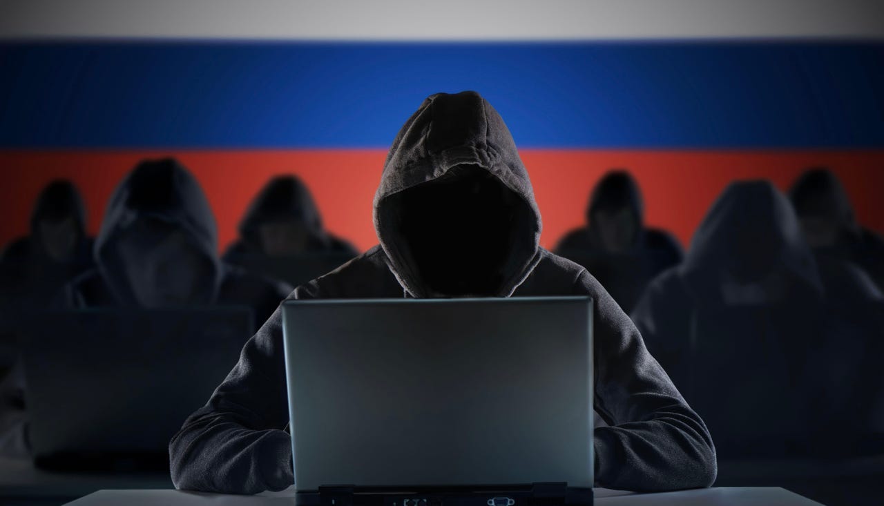 Seven hooded figured behind laptops in front of a background of the Russian flag