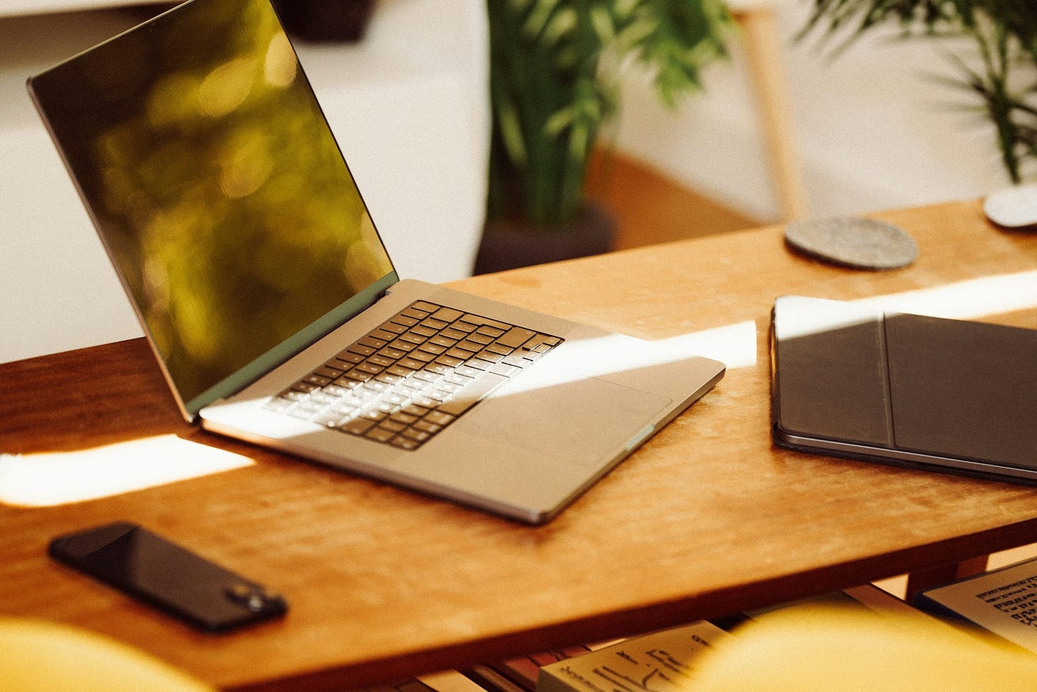 An open laptop, phone, and tablet sit on a wooden table with plants in the background