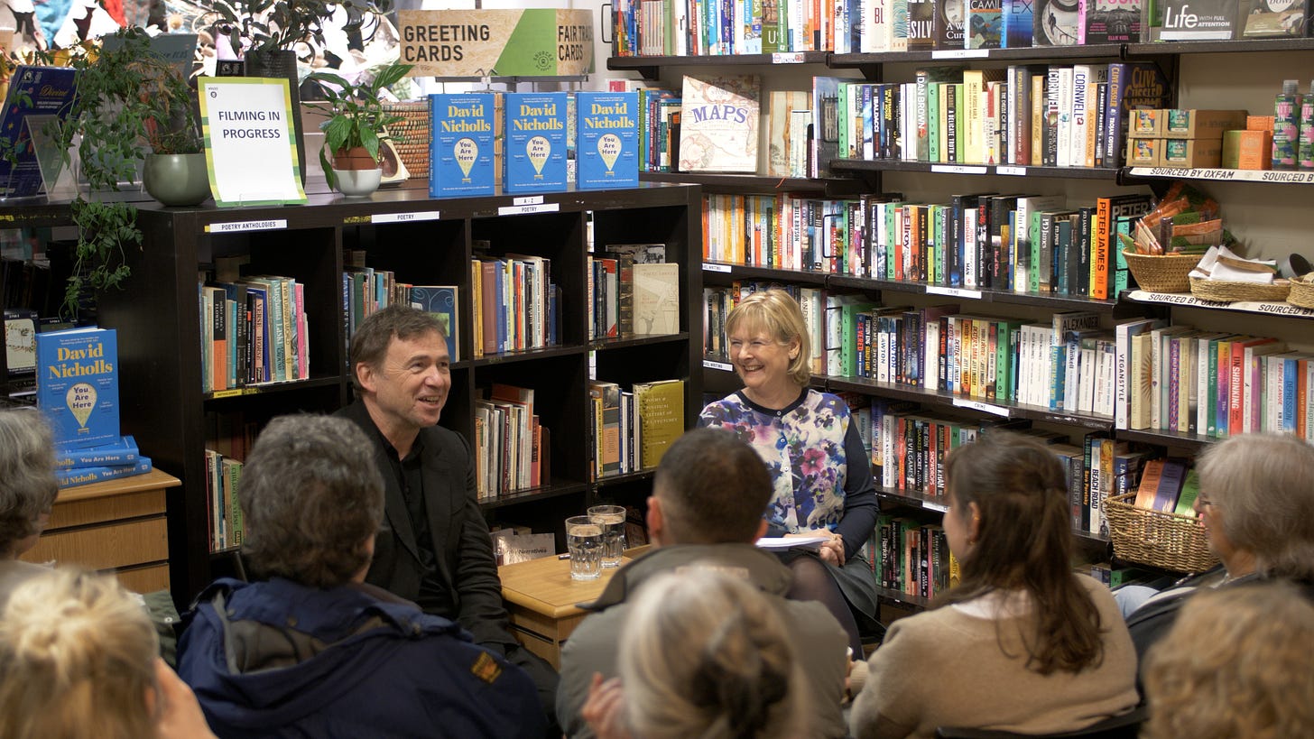 David Nicholls smiles while talking to Martha Kearney in Oxfam Books and Music Islington while seated in front of an audience. Several copies of his book, You Are Here, are visible behind him on a bookshelf.