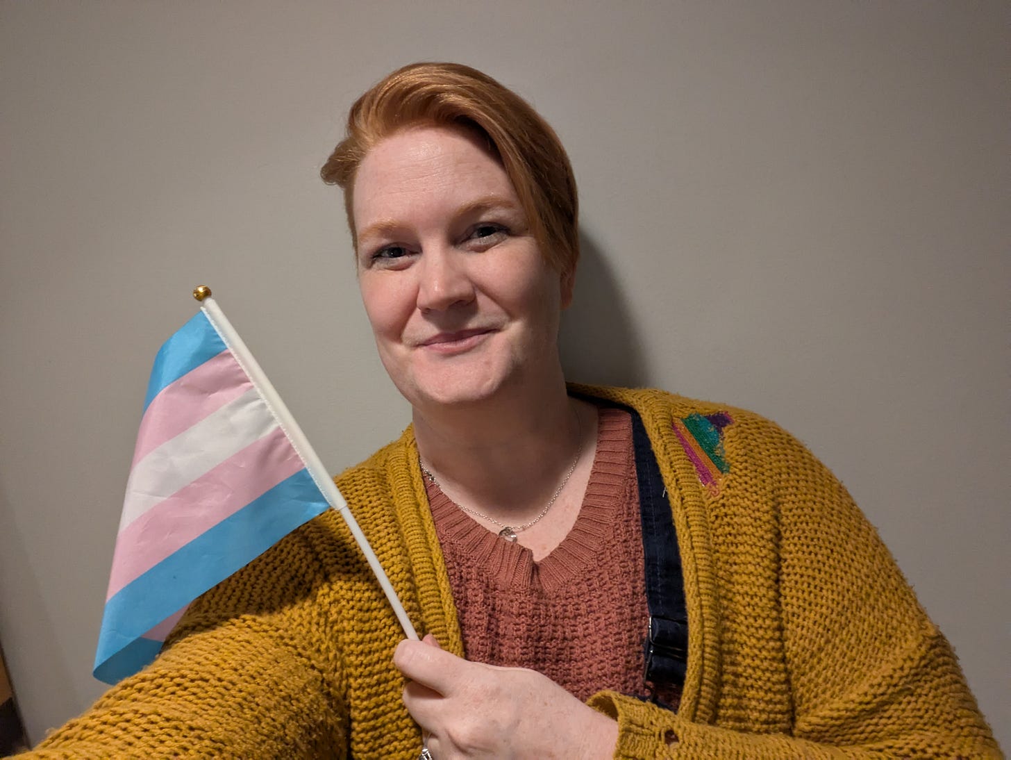 A photo of the same person as the first in this article, ten years later, wearing the same sweater--now patched with a rainbow--and holding a trans flag while smiling at the camera.