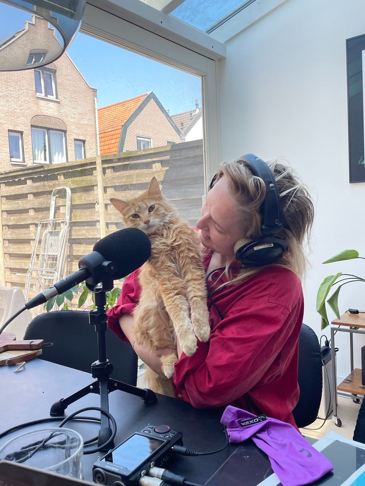 foto van Pleun die aan tafel op een stoel met koptelfoon op haar rode kat vasthoudt die voor de microfoon hangt en de camera in kijkt. Pleun heeft blond haar en draagt een rood shirt