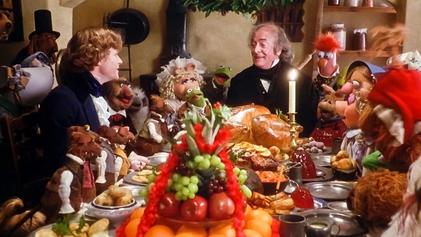 Scene from The Muppet Christmas Carol showing human actors and Muppet characters gathered around a lavish holiday feast table. The table is laden with a roasted bird, fruits, vegetables, and various dishes. Characters including Kermit the Frog, Miss Piggy, and others are dressed in Victorian-era clothing, depicting a festive celebration dinner scene.