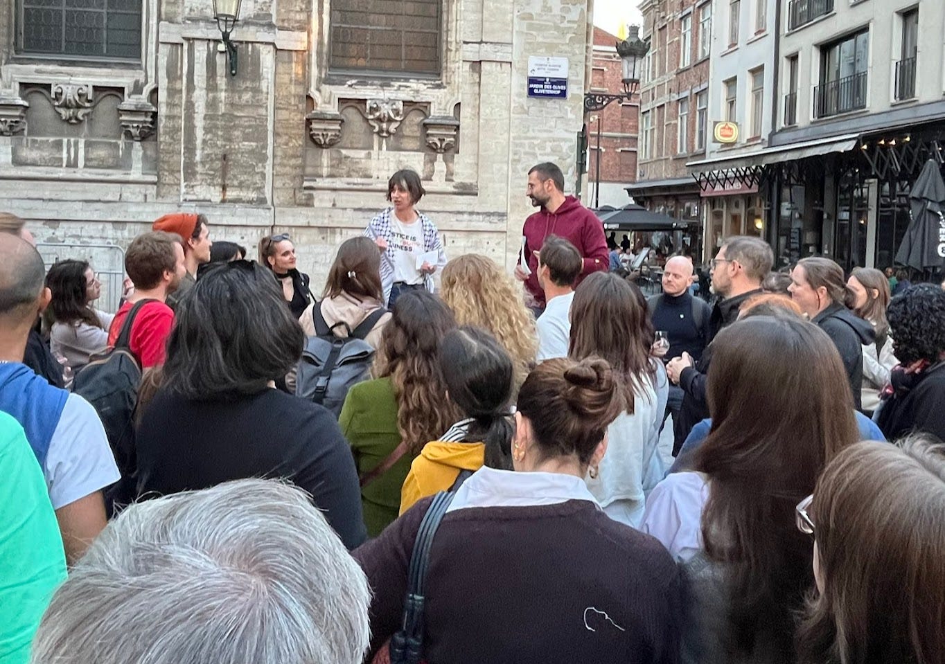 Cass and a white man stand on chairs in front of a crowd gathered outside, in front of a church and bars. Cass is wearing a keffieyh, white shirt and jeans and addressing the crowd while Alban, the man beside her, listens.