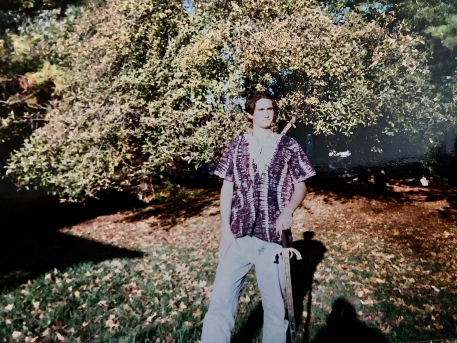 Dominic stands in a yard strewn with leaves wearing a purple and white dashiki and holding a sword like a cane Dominic stands in a yard strewn with leaves wearing a purple and white dashiki and holding a sword like a cane