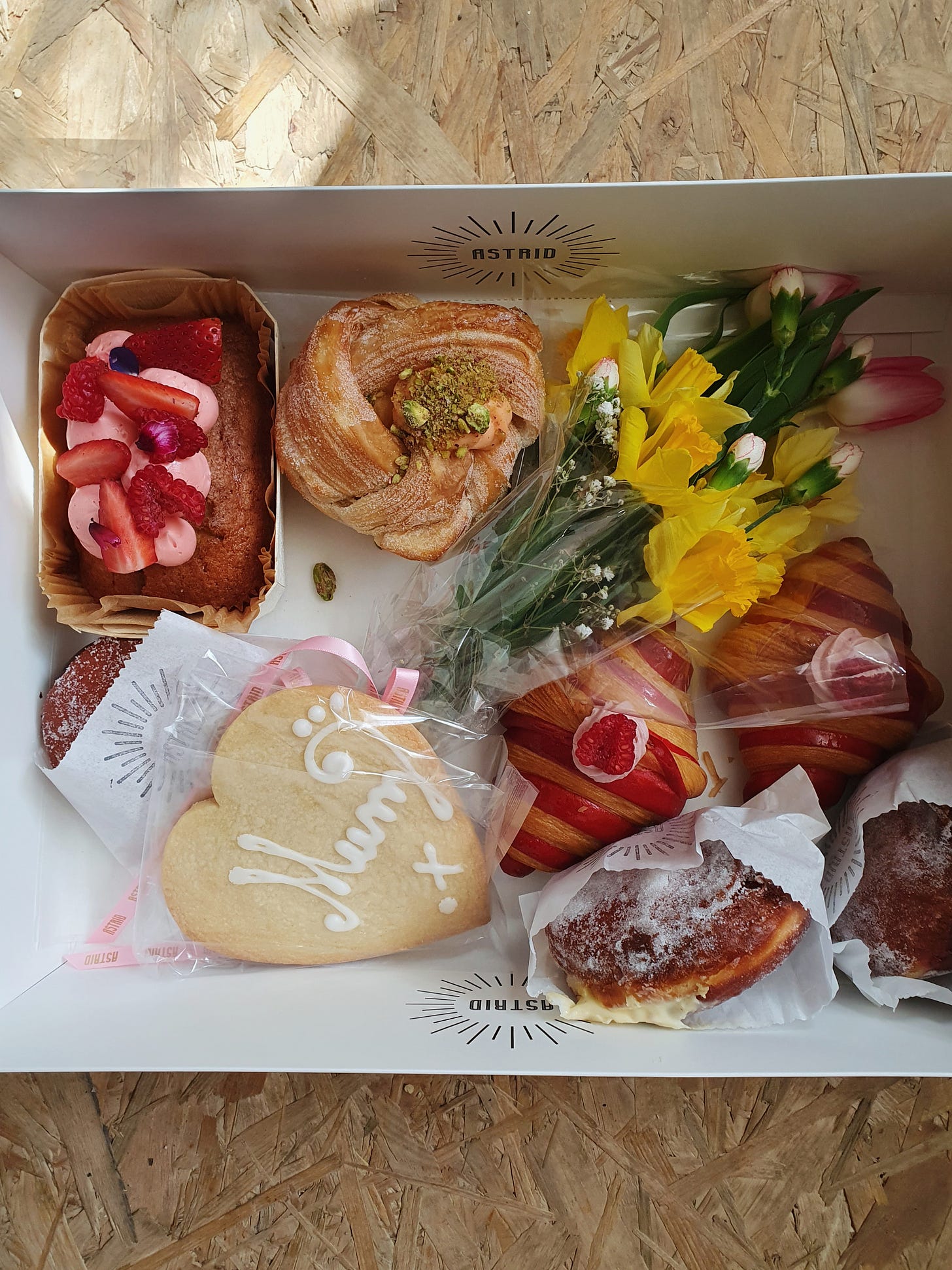 A picture of a box with a small bunch of flowers and lots of baked sweet treats including a heart shaped biscuit with the word mum iced on it.