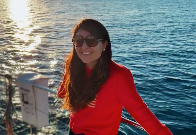 Woman in orange jumper and large sunglasses smiling with sea in the background