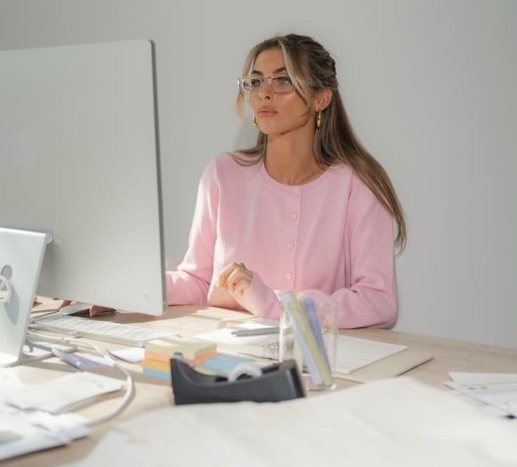 This may contain: a woman sitting at a desk in front of a computer monitor and keyboard, looking off to the side