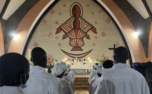 From behind, men and women in white robes line up in a church with candles, a large figurative mural of a black Christ on the wall in front of them. From behind, men and women in white robes line up in a church with candles, a large figurative mural of a black Christ on the wall in front of them.