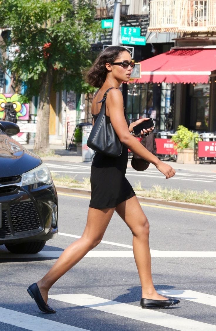 This may contain: a woman walking across the street with her purse in hand and wearing black shoes on