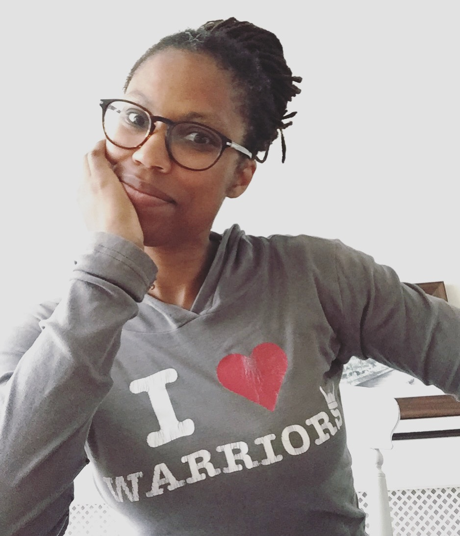 sandra wears glasses, her black dreadlocs pulled back with her favourite shirt that says "I (red heart) warriers" on it