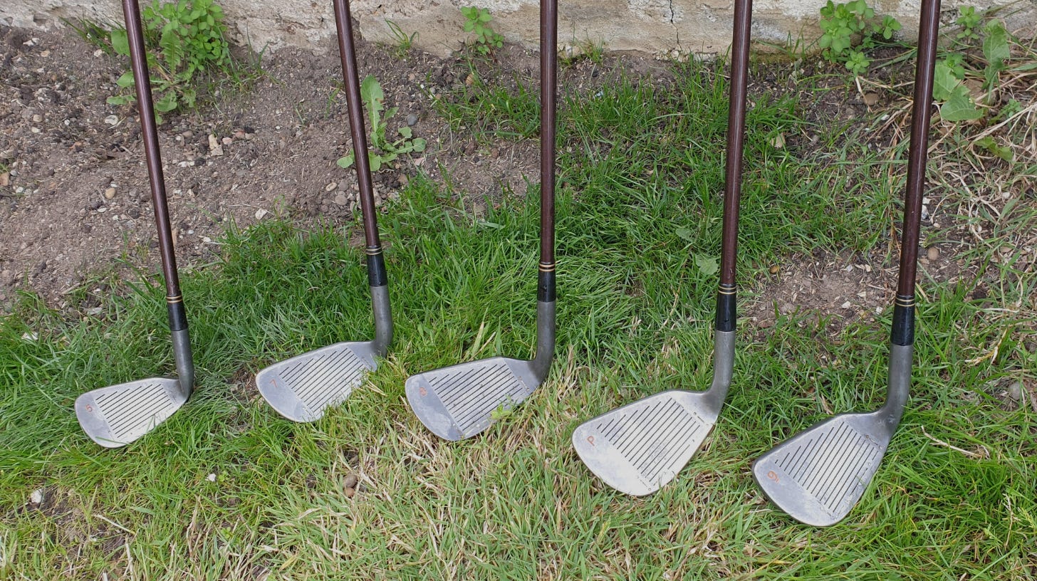 Six old golf irons with brown shafts lined up on grass