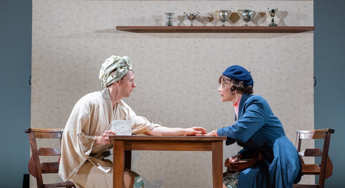 A man wearing a silk gown and hair wrap, and a woman wearing a long navy coat and matching hat sit, opposite one another at a wooden dining table. They are staring intently at each other, with the man reaching out one arm across the table in an attempt to reach the woman’s hand.
